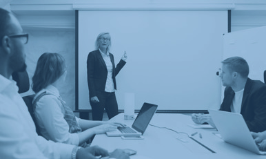 Tre personer vid ett bord lyssnar på en fjärde som talar vid en vit tavla.