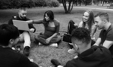 Sex personer sitter i ring på gräsmatta och diskuterar något som visas på laptop.