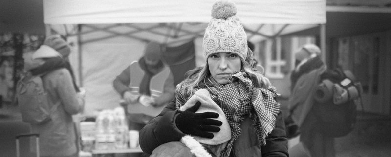 Ukrainsk kvinna står vid ett tält där vatten delas ut. Hon tittar allvarligt in i kameran medan hennes barn ledset trycker pannan mot kvinnan. Kvinnan omfamnar barnet.